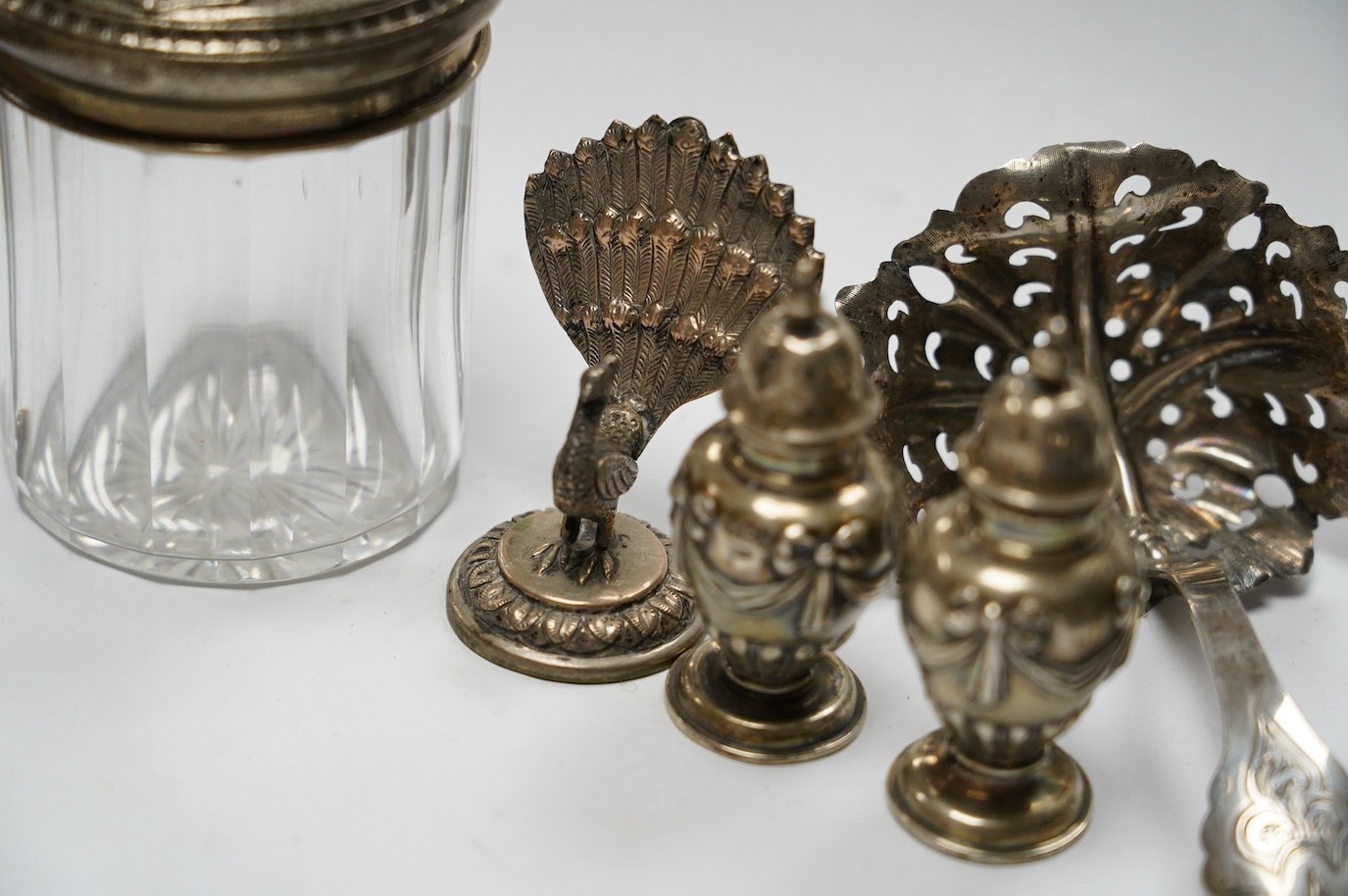 Three assorted modern silver wine labels, a ceramic wine label, a pair of silver pepperettes, a silver topped glass toilet jar, a Dutch white metal sifter spoon and seven plated items including a pair of peacock menu hol